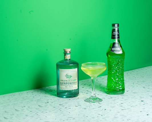 A bottle of Drumshanbo Citrus Gin, a green cocktail, and a bottle of Midori on a green background.