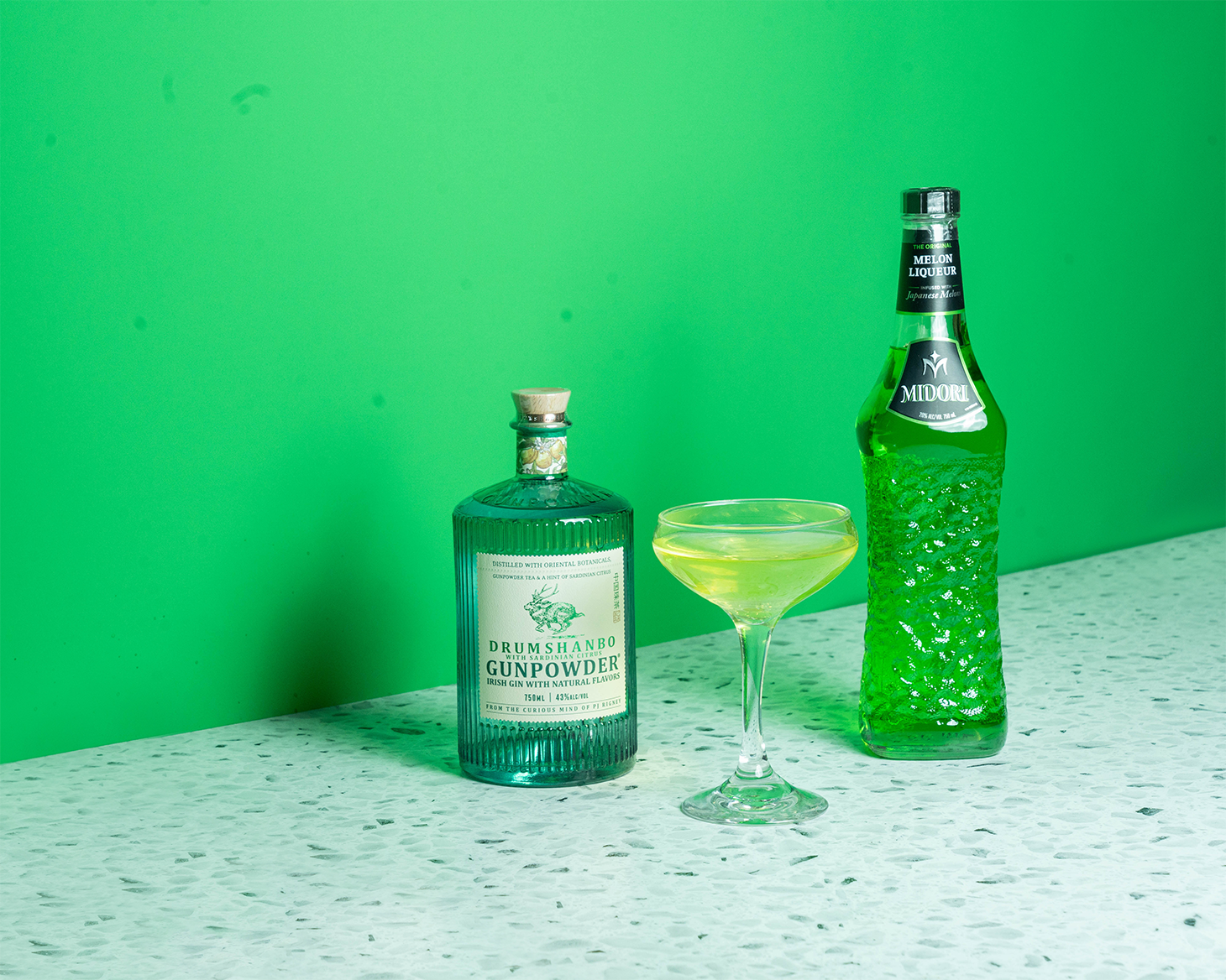 A bottle of Drumshanbo Irish Gin, a bottle of Midori and a green cocktail on a green background.