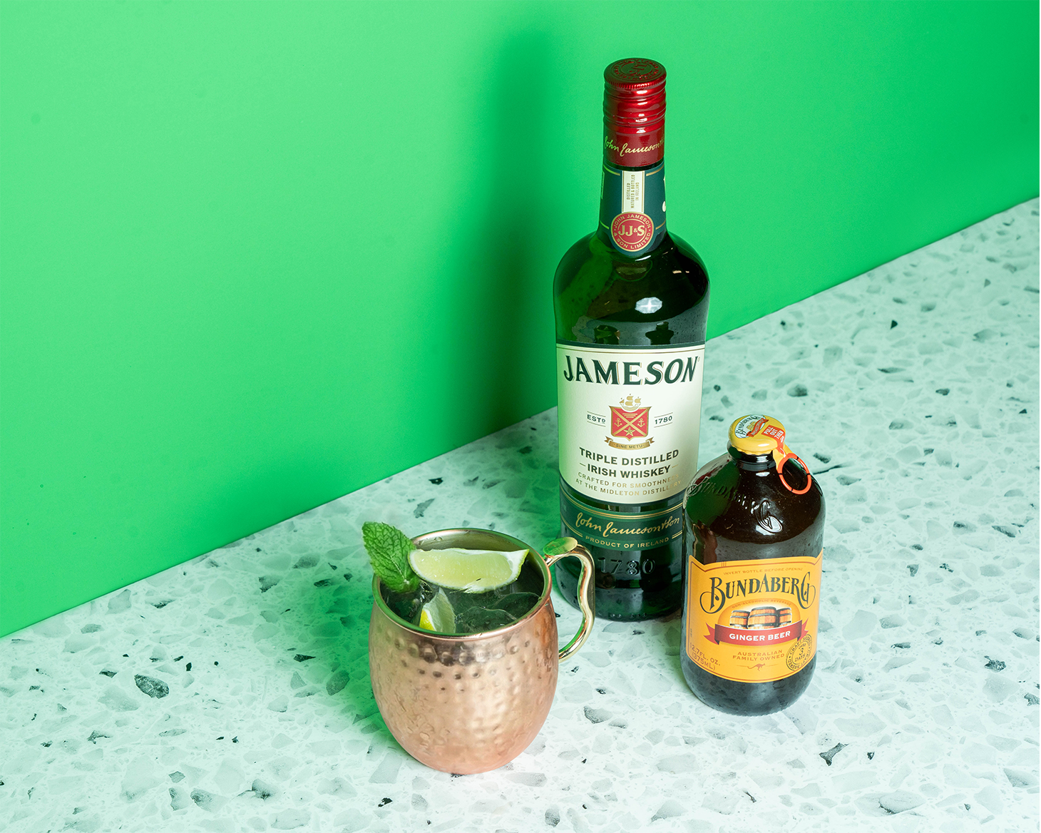A bottle of Jameson, a bottle of Bundaberg ginger beer, and a cocktail in a copper mug on a green background.